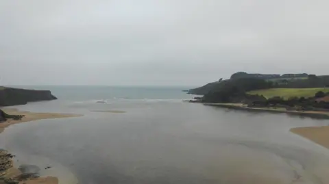The picture shows a wide estuary opening out towards the sea on a grey, overcast day. Shallow water spreads across the centre, with smooth sandbanks curving along both sides. The land on the right rises into rolling fields and patches of woodland, while the left side is edged by darker, rockier cliffs. Gentle waves break at the mouth of the estuary, where the inlet meets open water.