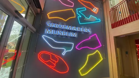 A neon sign on a wall inside a building reads Shoemakers Museum with drawings of shoes lit up around it. There is a glass door to the left of the image.