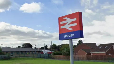 A sign for Horden railway station in a carpark. The sign is red with the white symbol of National Rail on it and says Horden.