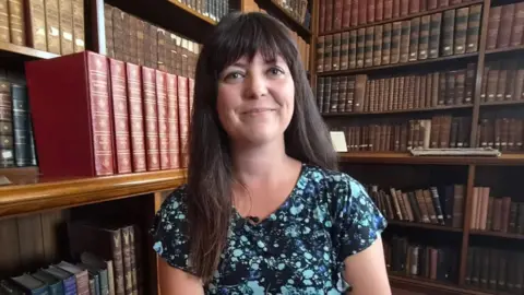 Helen Jenner is sat surrounded by books on shelves, with a large stack of red old-style books to the left of her head.