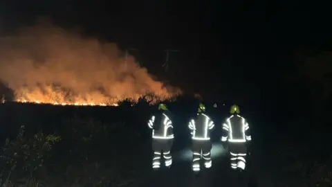 Mullion Community Fire Station Three firefighters walking towards a blaze in the dark. Their uniforms are being lit by the flash of a camera. 