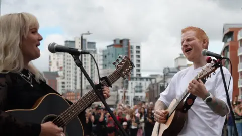 Shaun Whitmore/BBC A man with red hair and a white T-shirt plays a guitar alongside a woman with blonde hair and a black top.