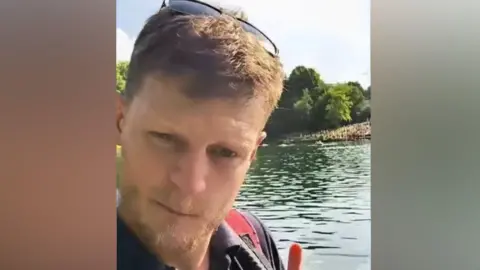 Facebook A man with short brown hair and a trimmed beard stands near a calm lake surrounded by trees. He wears a dark shirt, sunglasses on his head, and a red backpack strap over his shoulder, holding up one finger while looking at the camera under bright daylight.