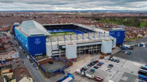 PA Media An aerial view image captures Goodison Park from up high. There is a car park situated at the front with housing surrounding the rest of stadium which is royal blue and white. 