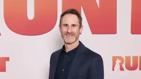 A grey-haired man wearing a dark blue suit smiles at the camera.