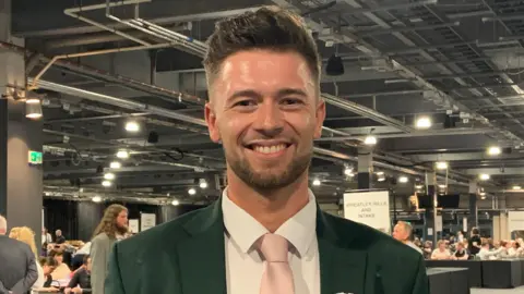 BBC/Lucy Ashton A man in a green suit and pink tie looks straight at the camera and smiles. It appears he is standing in a big hall with several people seated around tables in the background. A banner over his right shoulder reads "Wheatley Hills and Intake".