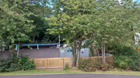 United Utilities View of a line of trees in front of a municipal type structure, with metal walls and a corrugated iron roof, behind a wooden fence. There is low shrubbery and a grassed area in front of the fence and more trees behind.