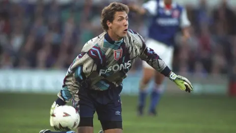Clive Brunskill/Allsport/Getty Images Ipswich Town goalkeeper Craig Forrest in action during a match against Leeds United at Portman Road. He is wearing goalkeeping gloves a long-sleeved top and navy Umbro shorts. He is in the process of throwing a football in an underarm motion.
