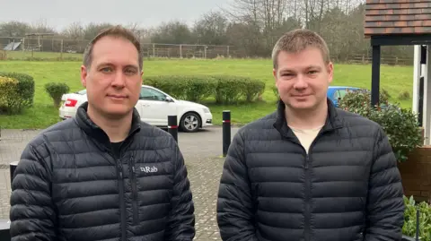 BBC Matthew and Anthony stood in front of the proposed development site. They're both wearing black puffer jackets and smiling slightly. Behind them is a green field with some hedges dividing it from the road. To Anthony's left is the porch of a house, and two cars - one white, one blue - can be see parked on the verge. 