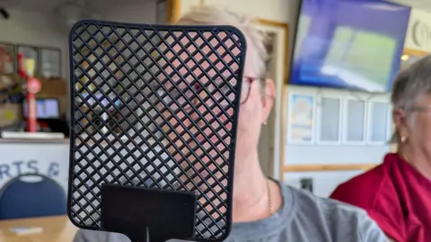 BBC A white lady with short grey hair and gold earings wearing a grey T-shirt sitting in a sports clubhouse with a black fly swatter in front of her face 