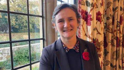 A woman, wearing a smart jacket and a poppy is smiling at the camera.