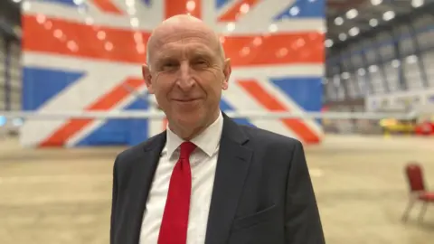 A head and shoulders shot of a bald man wearing a black suit jacket, white shirt and red tie smiling into the camera. A large union jack flag can be seen displayed behind him.