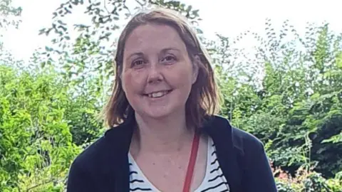 Linda-Jayne is smiling, wearing a fleece and low-cut top with horizintal stripes. Behind her is green shrubbery and across her chest is a handbag strap. 