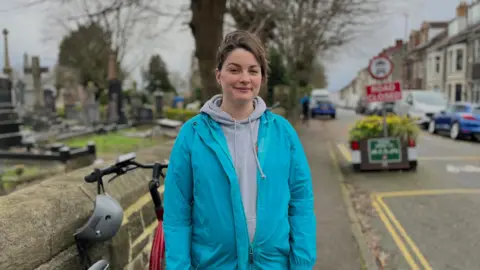 Lucy Husskison is wearing a bright blue coat and a grey hoodie underneath. Next to her is a bike leaning on a stone wall, separating the pavement of a residential street and a cemetery. Behind her left shoulder is a bollard and planter from the scheme. 
