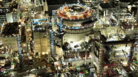 A very large nuclear power station under construction is lit up at night. There is a large dome at the top of the largest part of the site, and there is scaffolding everywhere.