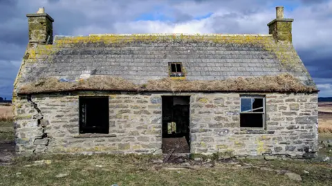 Angus Mackay Photography The cottage has a big crack down one exterior wall. There is no front door or glass in the windows. Yellow lichen covers the slate roof.