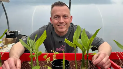 Alastair Hawken is crouching in front of a red plant container which contains several ginger plants.  He is standing in the polytunnel where the ginger is being grown.
