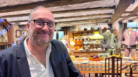 Lily-May Symonds/BBC Gary Tomkins stands in front of the Burrow set at the Harry Potter Studio Tour in Watford, with a rustic kitchen table, hanging lamps, and mannequin costumes visible behind them. Gary is wearing a navy blue jacket and white shirt. He has black glasses and grey hair. 
