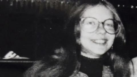 A black and white photograph of a smiling woman wearing glasses with long hair