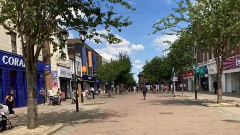 An image of a pedestrianised street in Rotherham town centre.  The road is paved and tree-lined, with shops on either side. People can be seen walking along the pavement, some pushing prams.