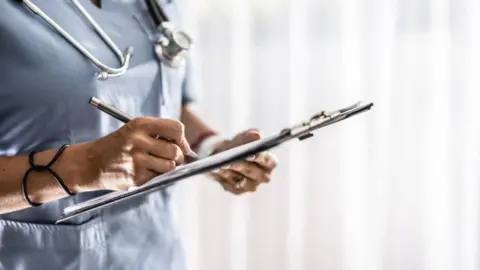A nurse in blue uniform, holding a clipboard and writing on it. There is a stethoscope around their neck.
