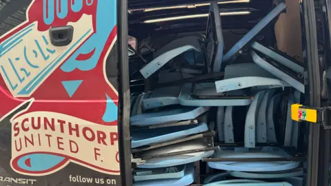 Julie Taylor The back of a van with one door open and blue seats packed inside. The other door is closed and decorated with a large transfer showing the Scunthorpe United club logo - a claret and blue fist hold an iron bar, with the text "Scunthorpe United FC".