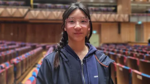 A teenager is standing in an auditorium with chairs around her in the background. She has long, dark hair and is looking at the camera.
