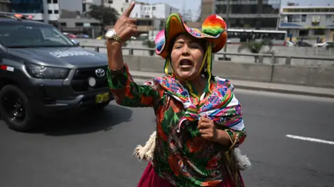 Getty Images A traditional woman wearing a colorful hat and clothes raises her index finger and shouts against former president Fujimori. A photo of a Toyota car in the background