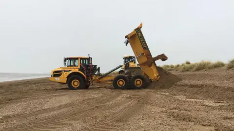 Environment Agency Yellow diggers transporting sand across the beach.