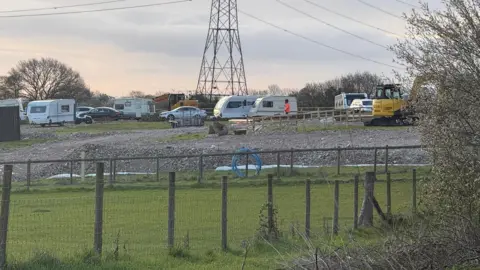 A view of the field. About six caravans are parked on hardstanding along with other vehicles and yellow diggers. There is a pylon in the middle of the field.