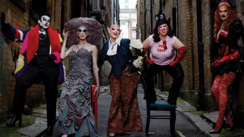 Magnus Hastings Five drag performers pose in an alley. The first, dressed as a man wears a white face and multi-coloured coat. The second has a grey dress with a large round hate. The third wears a blazer and wide trouers, the third stands on a chair in a t-shirt and red elbow-length gloves. The last rests with her back on the wall wearing red patterned tights and a corset.