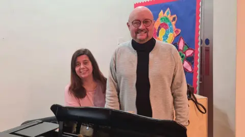 Ryan Longridge and Sarah-Jane Cooper by the communal grand piano in 2 Royal Avenue.  Sarah-Jane has long, dark hair and is wearing a pink top.  Ryan is bald with a goatee beard.  He is wearing glasses, a black top and beige cardigan.