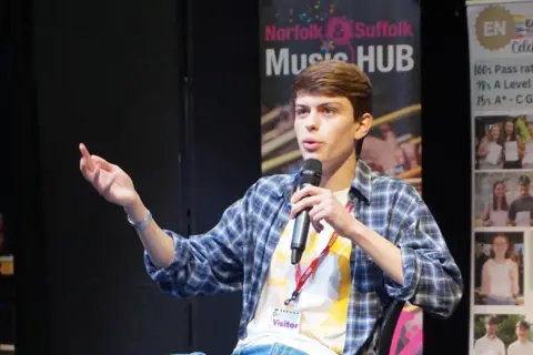 Norfolk County Council Tom Major sitting on a chair on a stage with a microphone in his left hand.