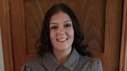 Darcy Ford Darcy Ford has long, dark brown hair and is smiling at the camera. She is wearing a checked top with a collar and is standing in front of a wooden door