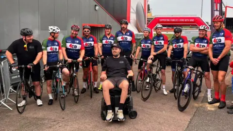 Gloucester Rugby Eleven people sitting on their bikes in a line, with their legs either side. They are all wearing blue, red and white cycling shirts that say '4Ed' on them. Ed Slater is sitting in his wheelchair just in front of them 