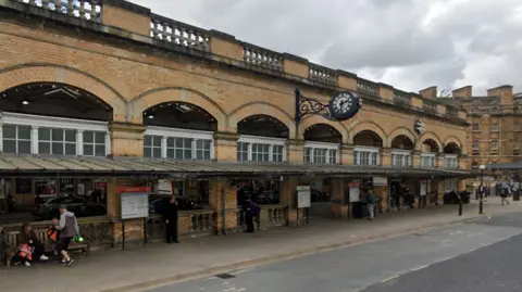 A long strain station building with an awning over the front and a clock above the shelter. Some people are sat on a bench. A bus stop is marked on the road in front of the building.