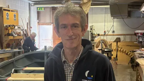 A man stands in front of the grey vessel in a workshop , all around are woodworking tools 