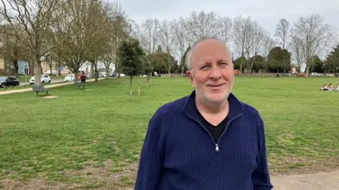 Henry Godfrey-Evans/BBC A man in a blue cardigan smiling with grass in the background