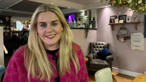 A woman with long blonde hair and a pink coat is sitting in a cafe area. She is smiling. Behind her there are chairs and tables and some greenery. There are also sleeves of pictures and framed photographs. 