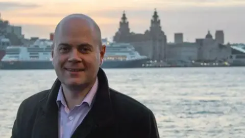 Handout Liverpool council leader Liam Robinson standing on the waterfront in Wirral, with Liverpool's Three Graces visible across the Mersey behind him