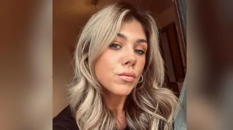 A woman with long blonde hair looks into the camera as she takes a selfie. She has green eyes and wears red lipstick. She has a silver hoop earring in her left ear. 