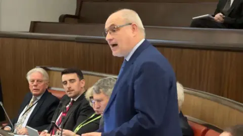 Adrian Warwick addresses the full meeting of Warwickshire County Council. He wears a blue suit while standing. Either side of him are councillors seated on red leather chairs with wooden panelling visible.