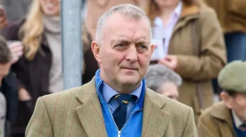Alan Raeburn A man with grey hair in a green tweed jacker with blue jumper, shirt and tie underneath. There are people out of focus in the background.