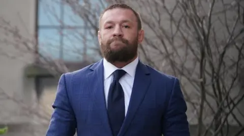 PA Media Conor McGregor walking into court. He is wearing a blue suit jacket with a white shirt and a navy tie.