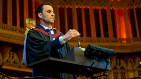 University of Bristol Mr Fattouh is photographed wearing a black robe with red and white accents, as well as a blue tie. He is standing in front of a dark brown plinth with a microphone.