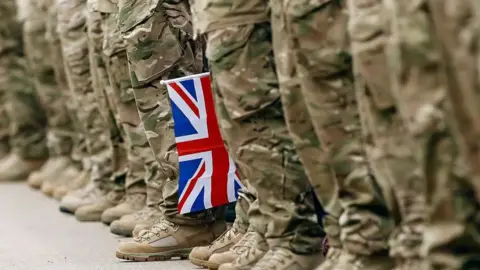 Boot-clad feet stood in a line with military-style trousers. A British flag sits between the legs.