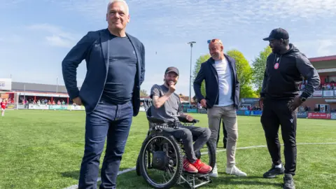 Jay Wrighte Four men are at the edge of a football pitch. Three are standing with one in a wheelchair.