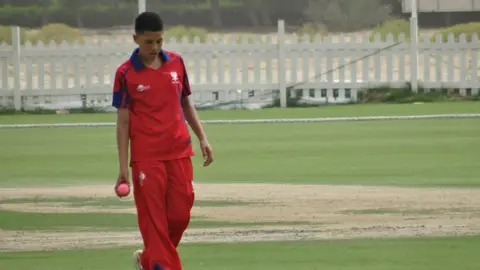 Bedford School Gay as a child, wearing a red cricket outfit, holding a ball in his left hand. He is walking on a cricket pitch. 