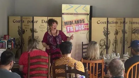 Poets, Prattlers and Pandemonialists The image shows a small live performance in an intimate indoor setting, where a performer stands and speaks in front of a seated audience. Behind the performer is a banner reading “Midlands Fringe,” indicating the event, while the audience sits closely together on chairs and sofas, watching the act.
