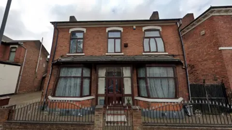 A two storey dethatched house surrounded by a brick and metal gate. It was long windows with brown wooden frames along the perimeter 